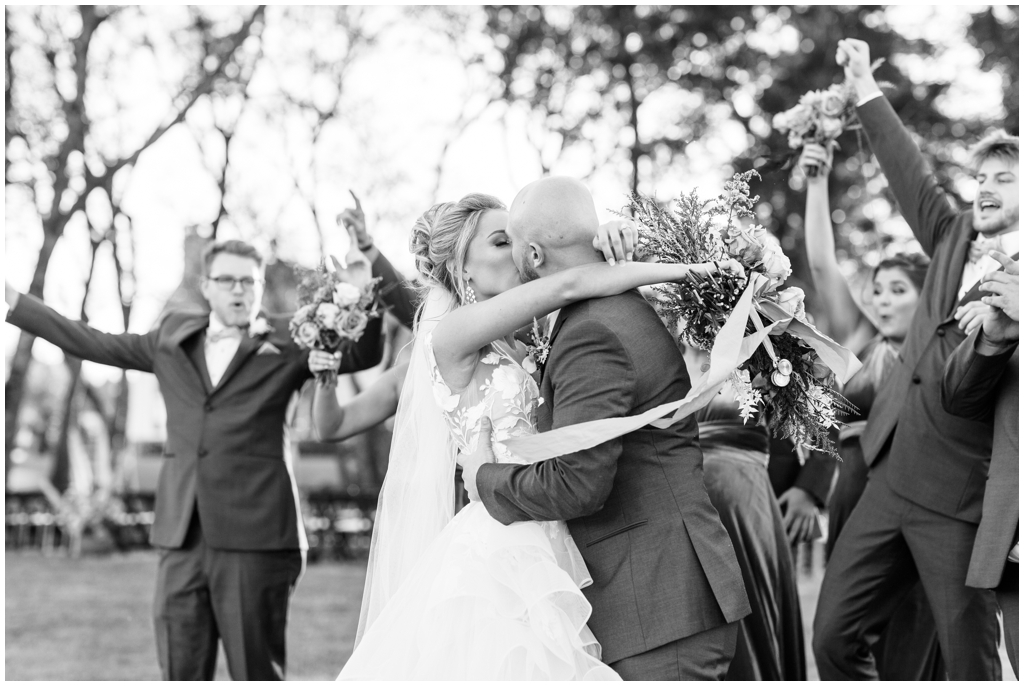 Bride and groom share a kiss surrounded by celebratory bridal party